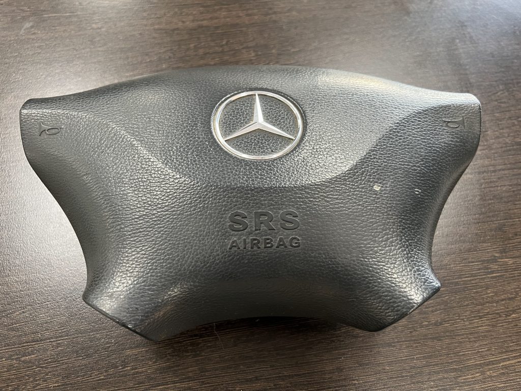 Close-up of a Mercedes-Benz steering wheel airbag module, featuring the iconic logo in the center and "SRS AIRBAG" text beneath it. The black surface has a textured finish, against a dark wooden background.