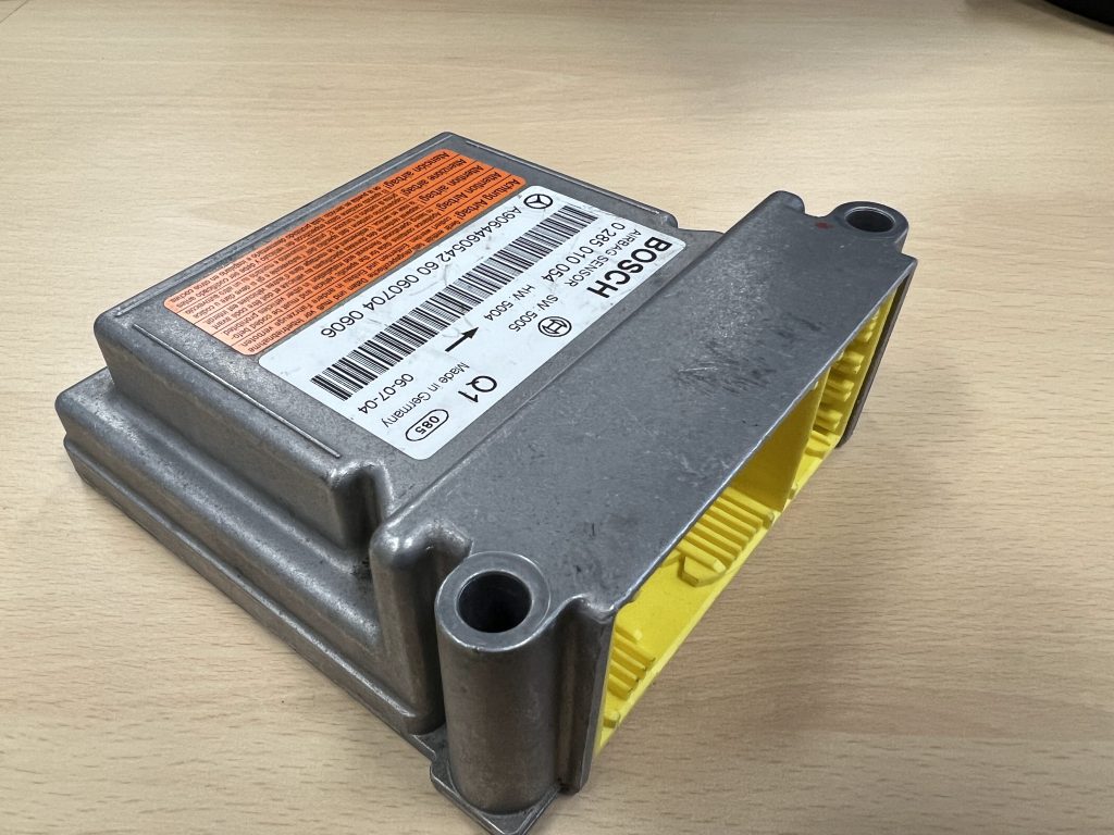 A silver metal automotive control module with a yellow connector on one side. The module, displaying a Bosch label and a prominent orange warning sticker is placed on a light wooden surface.