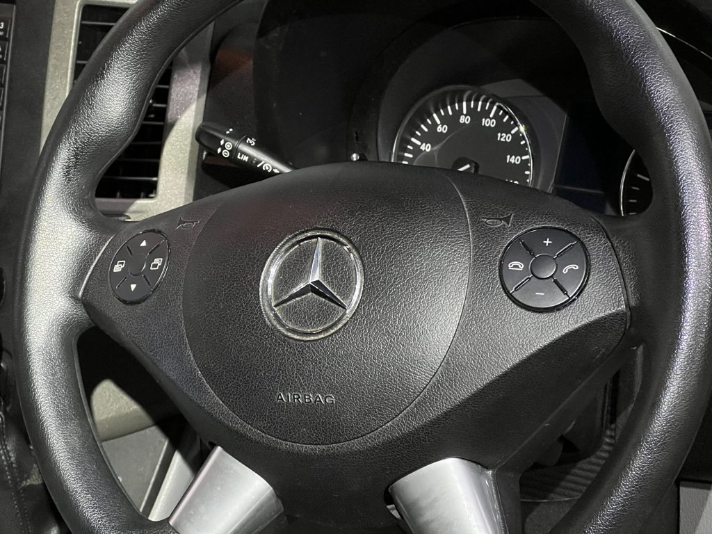 Close-up image of a Mercedes-Benz steering wheel. The wheel features the iconic Mercedes-Benz logo at its center, with control buttons on either side. Visible behind the wheel are part of the dashboard, including the speedometer with measurements up to 160 mph and a prominent SRS warning light indicator.