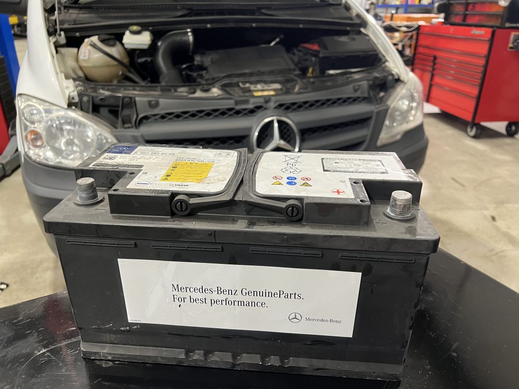 A close-up of a Mercedes-Benz car battery on a black surface in a garage. Behind it, the hood of a white Mercedes-Benz van is open, revealing the engine with a low battery sign. A red toolbox is partially visible on the right. The battery has a label stating "Mercedes-Benz Genuine Parts.