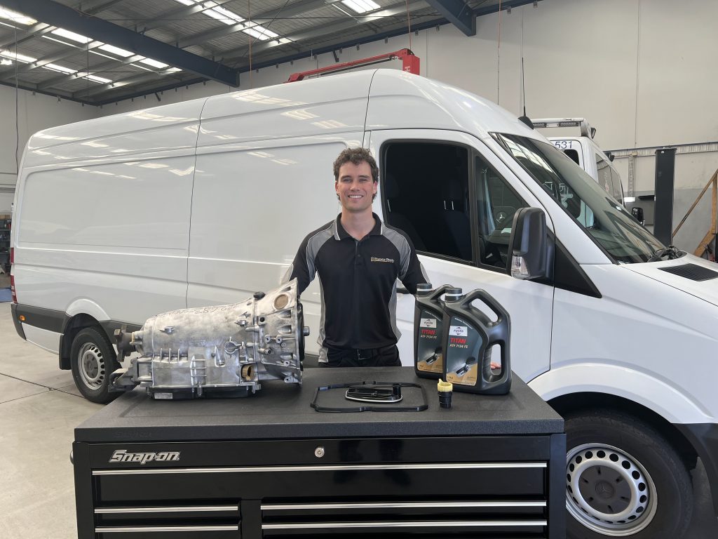 A man stands smiling in front of a white van in a garage. He's wearing a black polo shirt with a company logo. On the black workbench in front of him are an engine component, two containers of motor oil, and a wrench—ready for fuel injector repairs. The garage has tools and equipment around.