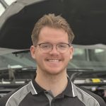 A person with short, light brown hair and glasses is smiling at the camera. They are wearing a black and gray collared shirt. In the background, there is an open car hood in a garage-like setting, perhaps indicating their readiness to help you make a booking for your next car service.