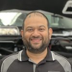 A man with short hair and a beard is smiling. He is wearing a black and gray collared shirt. The background appears to be a car with its hood open, suggesting he might be ready to help you out—don't hesitate to make a booking!