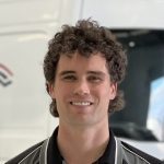 A smiling man with curly dark hair is standing indoors. He is wearing a black polo shirt with white piping details. The background, slightly blurred, features part of a vehicle and a building—a perfect snapshot before you make a booking for your next adventure.