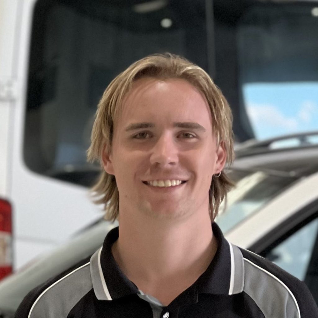 A man with shoulder-length blonde hair smiles at the camera. He is wearing a black and gray polo shirt. Behind him, there is a blurred background of a vehicle inside an indoor space, possibly a showroom or garage, hinting that it's the perfect place to make a booking for your dream car.