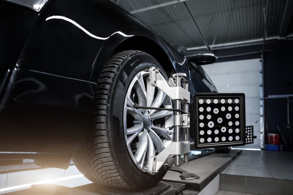 A close-up view of a car's wheel being aligned in an auto repair shop. The tire is secured with a specialized clamp, and a diagnostic device with a grid and dots is attached to the wheel. In the background, you can see equipment for various services like fuel injector repairs and a partially open door.