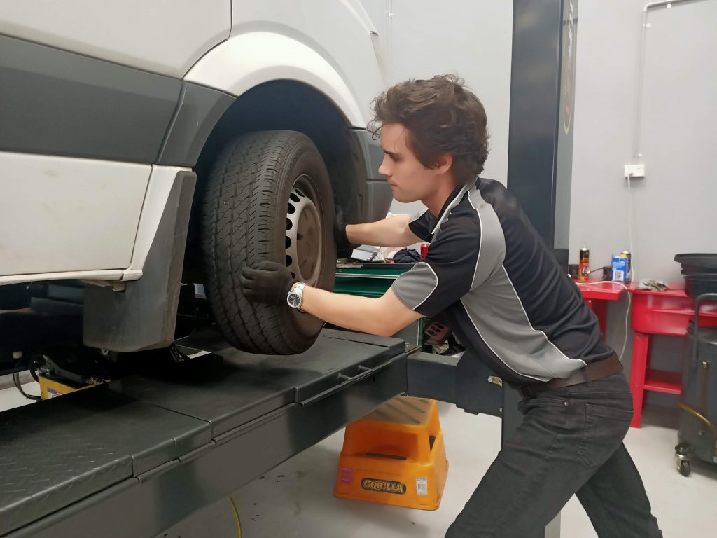 A mechanic wearing gloves is working on a white vehicle lifted on a platform. He is using both hands to attach or remove a tire, possibly before addressing fuel injector repairs. Tools and automotive equipment can be seen in the background.