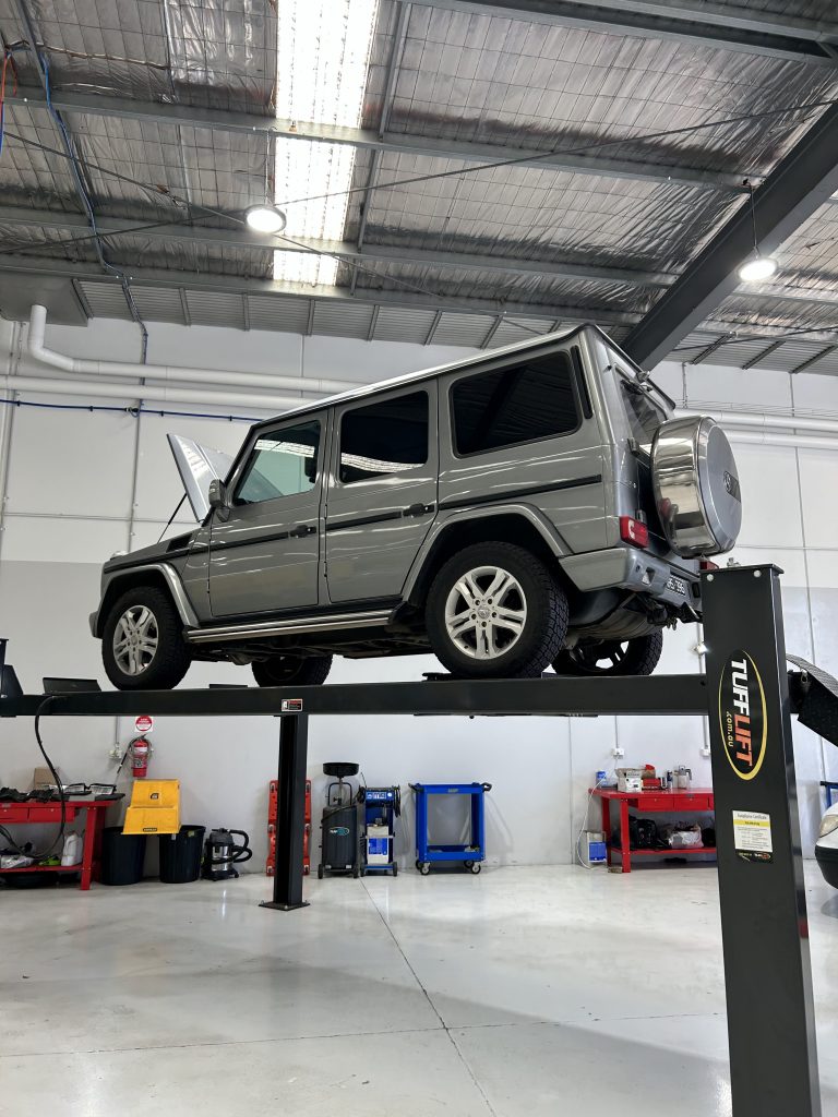 A gray luxury SUV is lifted on a hydraulic car lift in an automotive repair shop. The hood is open, and various tools and equipment are visible around the workshop. As the Mercedes undergoes maintenance or inspection, misconceptions of diesel engines often come to mind.