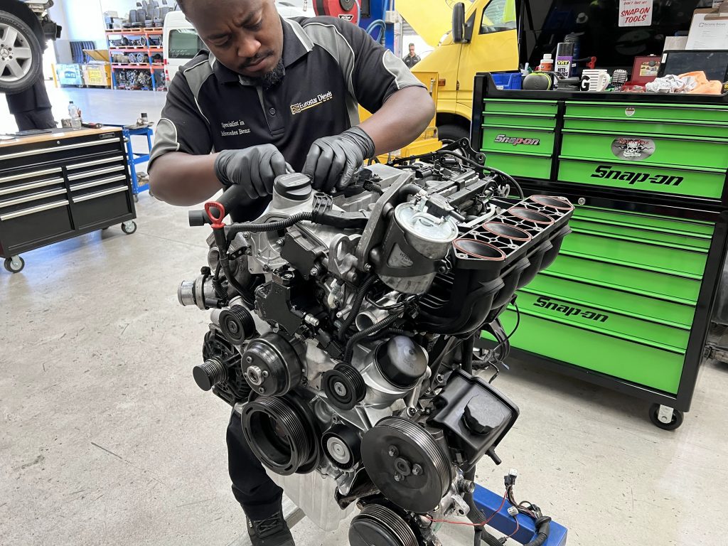 A mechanic specializing in wheel alignment is working on an engine in a garage. He is wearing a black and gray uniform with gloves, focusing intently on his task. The background features various tools, a tool chest, and equipment, including a bright green Snap-on tool cabinet.